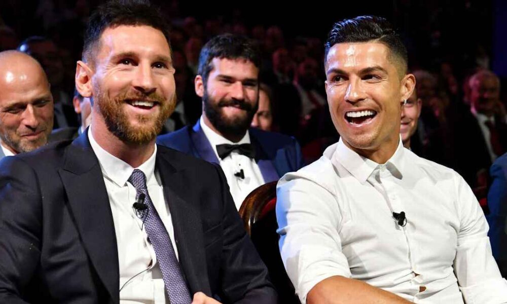 Messi e Cristiano Ronaldo sentados lado a lado e sorrindo durante sorteio dos grupos da Champions League temporada 2019/2020