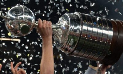 Close-up da taça da Libertadores da América, mostrando os detalhes em prata e a base de madeira com as placas dos clubes campeões.