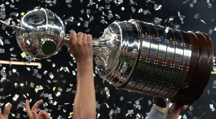 Close-up da taça da Libertadores da América, mostrando os detalhes em prata e a base de madeira com as placas dos clubes campeões.
